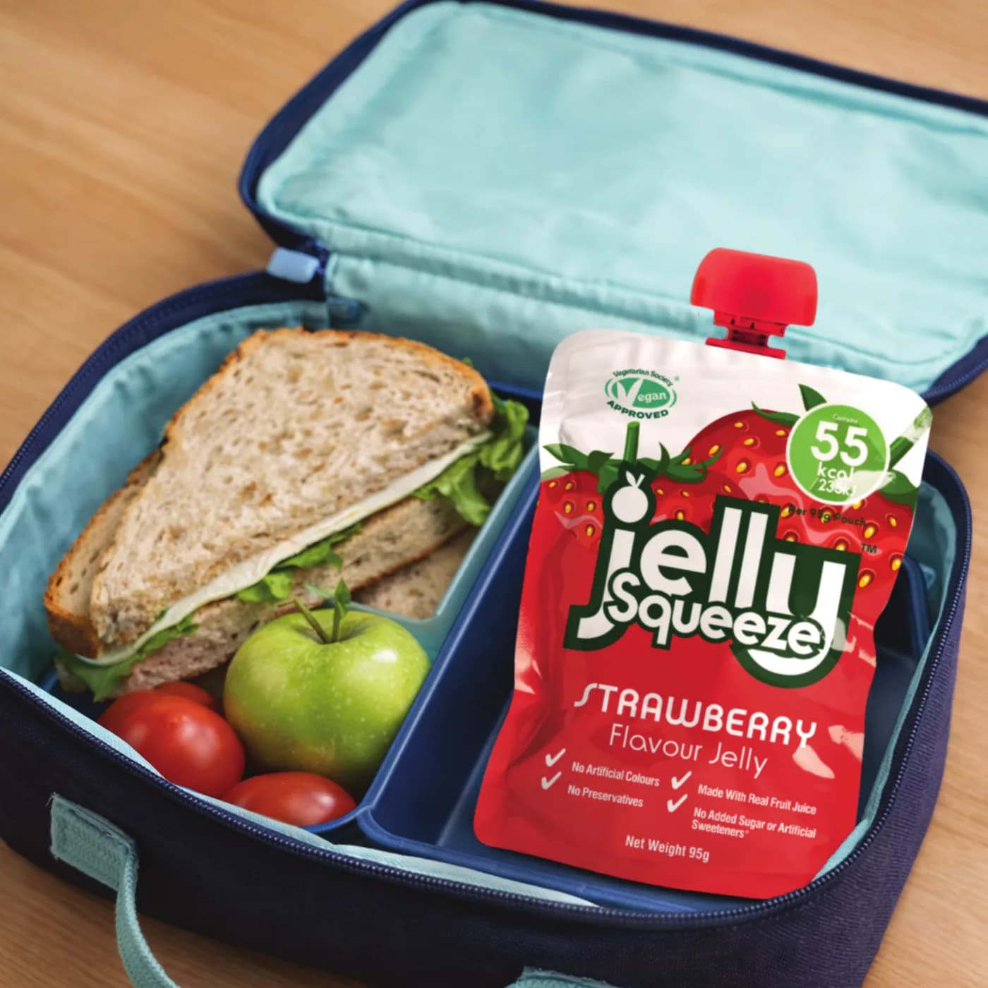 A Jelly Squeeze Strawberry pouch in a lunchbox next to a sandwich and an apple
