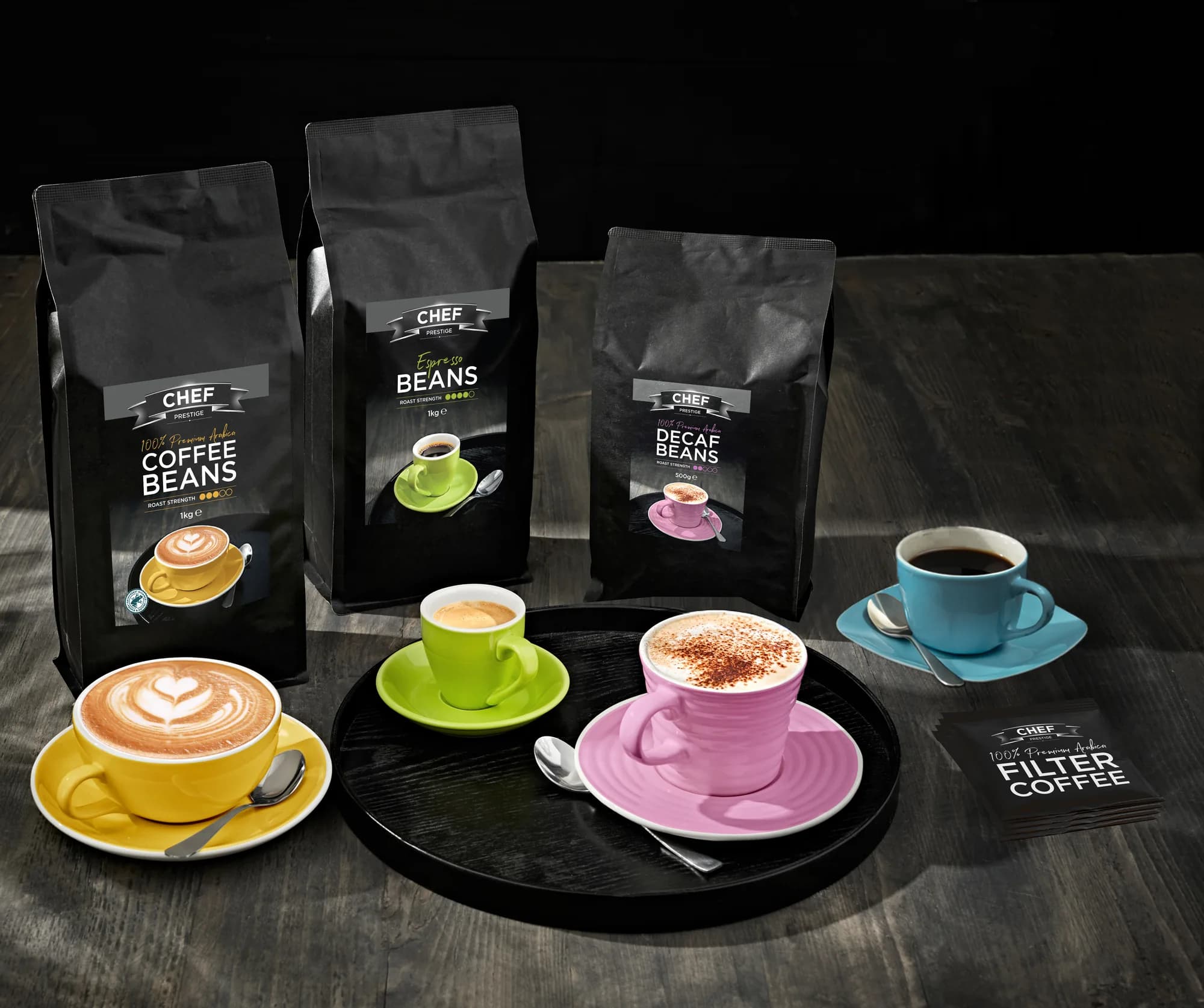 Chef Prestige Coffee Beans, Espresso Beans and Decaf Beans bags in matte black, with coloured cups on a café table